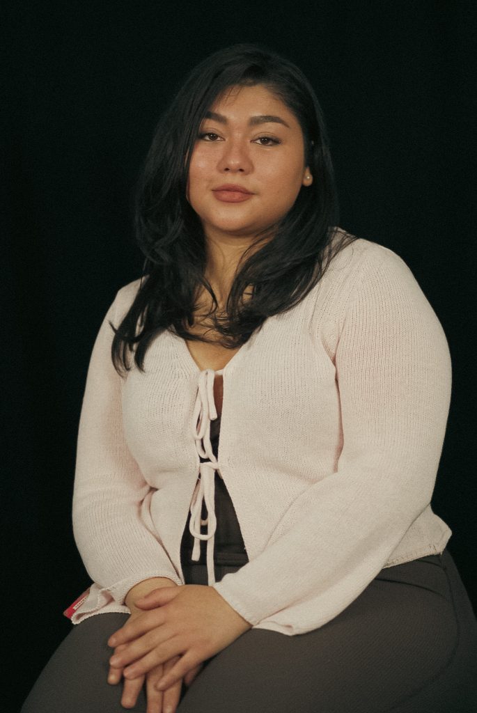 A woman with long dark hair wearing a light pink cardigan and dark pants sits with her hands folded in her lap against a black background. She looks calmly at the camera.
