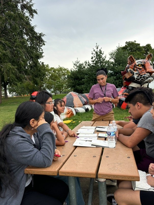 A group of young people sits around a picnic table outdoors, listening to a person in a purple shirt standing and speaking. Papers and water bottles are on the table, and tents and trees are in the background.