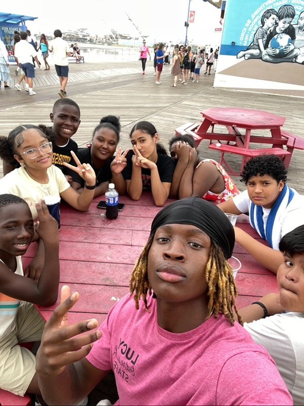 A group of eight smiling teenagers sits around a red picnic table outdoors, posing for a selfie and making peace signs. Other people and boardwalk scenery are visible in the background.