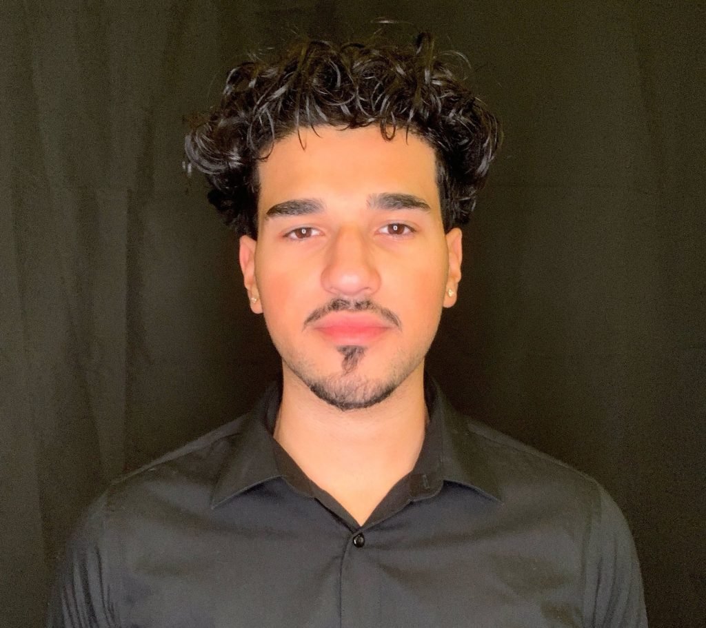 A young man with short, curly dark hair, trimmed facial hair, and earrings is wearing a black button-up shirt and standing against a plain black background, looking directly at the camera with a neutral expression.