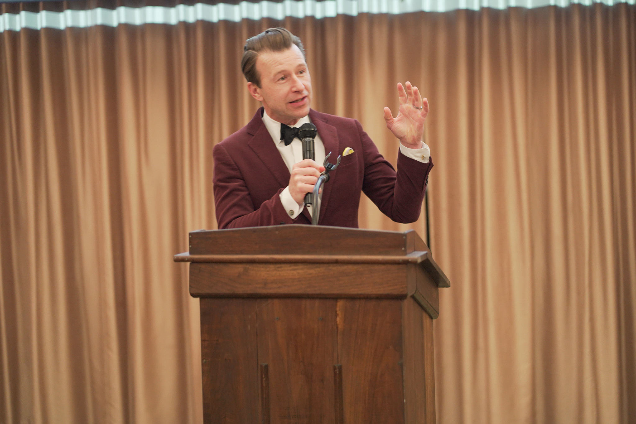A man in a maroon suit and bow tie speaks into a microphone while standing at a wooden podium, gesturing with his left hand, in front of beige curtains.