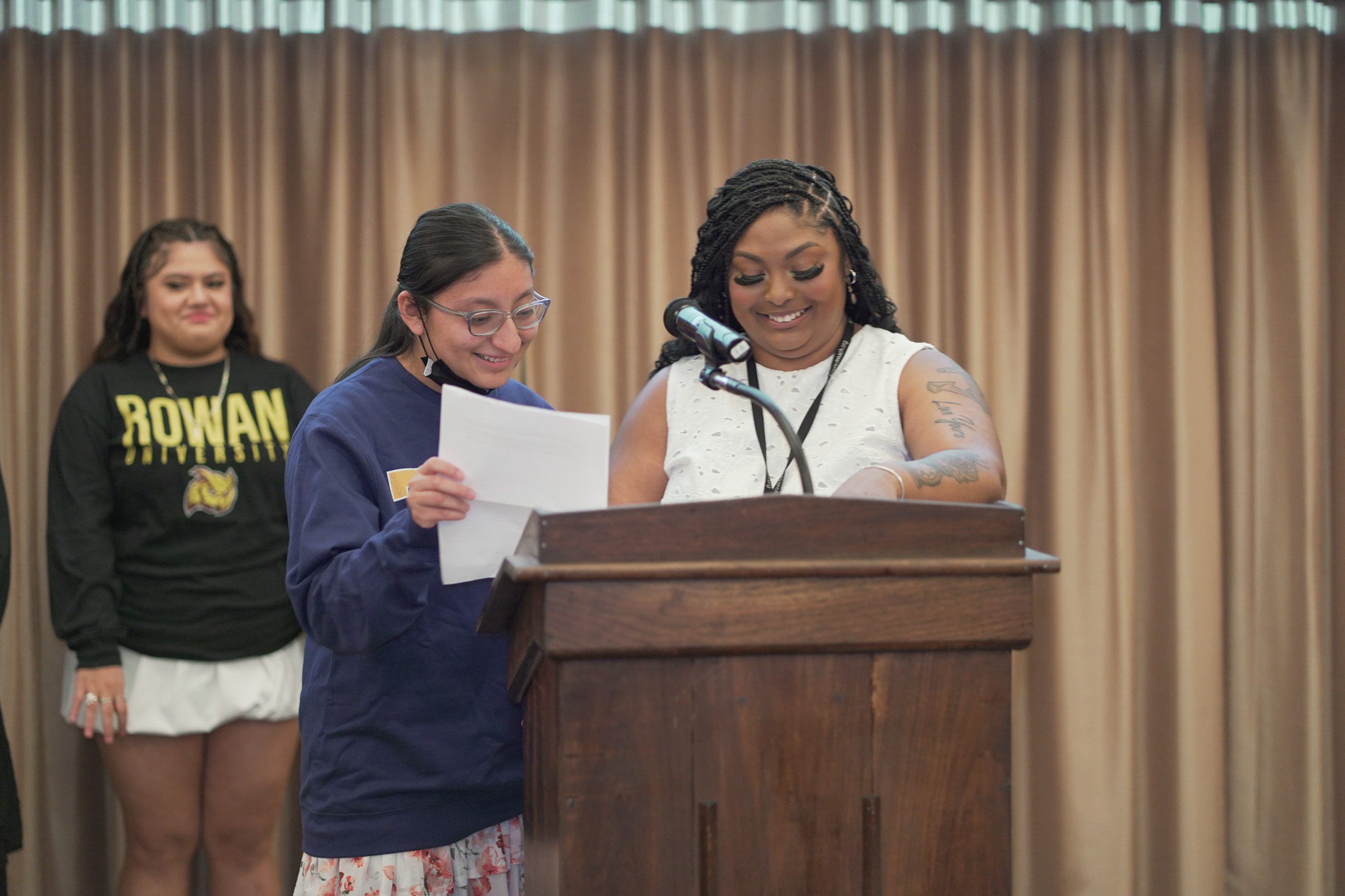 Two women stand at a podium smiling and holding papers, while another woman in a Rowan University shirt stands in the background. Light brown curtains hang behind them.