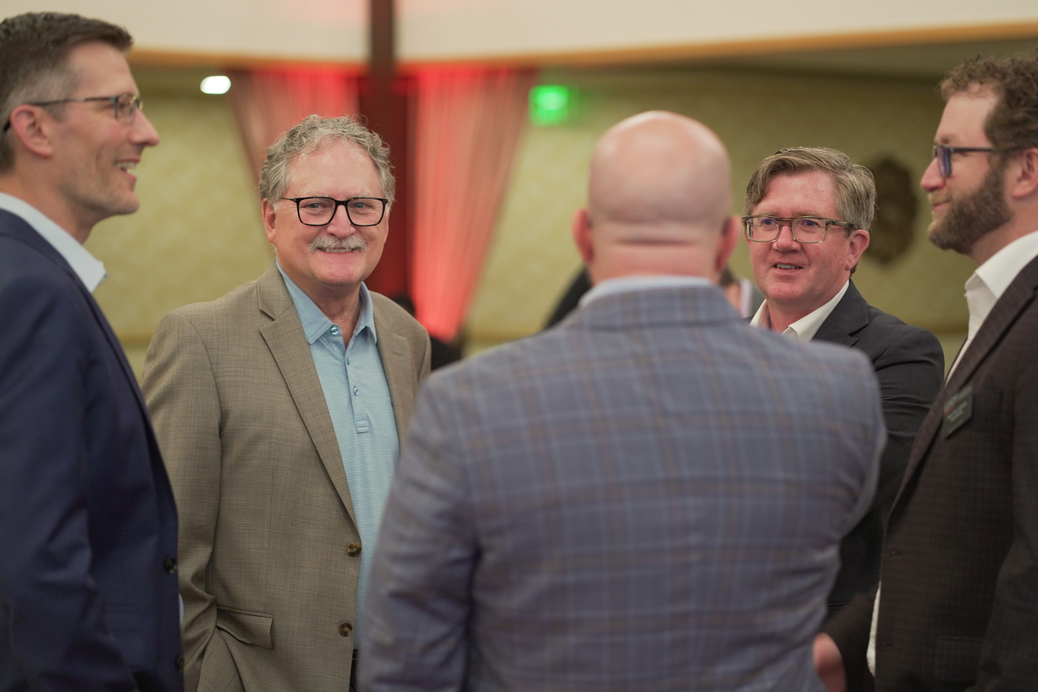 Five men in business attire stand in a circle indoors, smiling and conversing at what appears to be a formal or networking event, with warm lighting and blurred background décor.