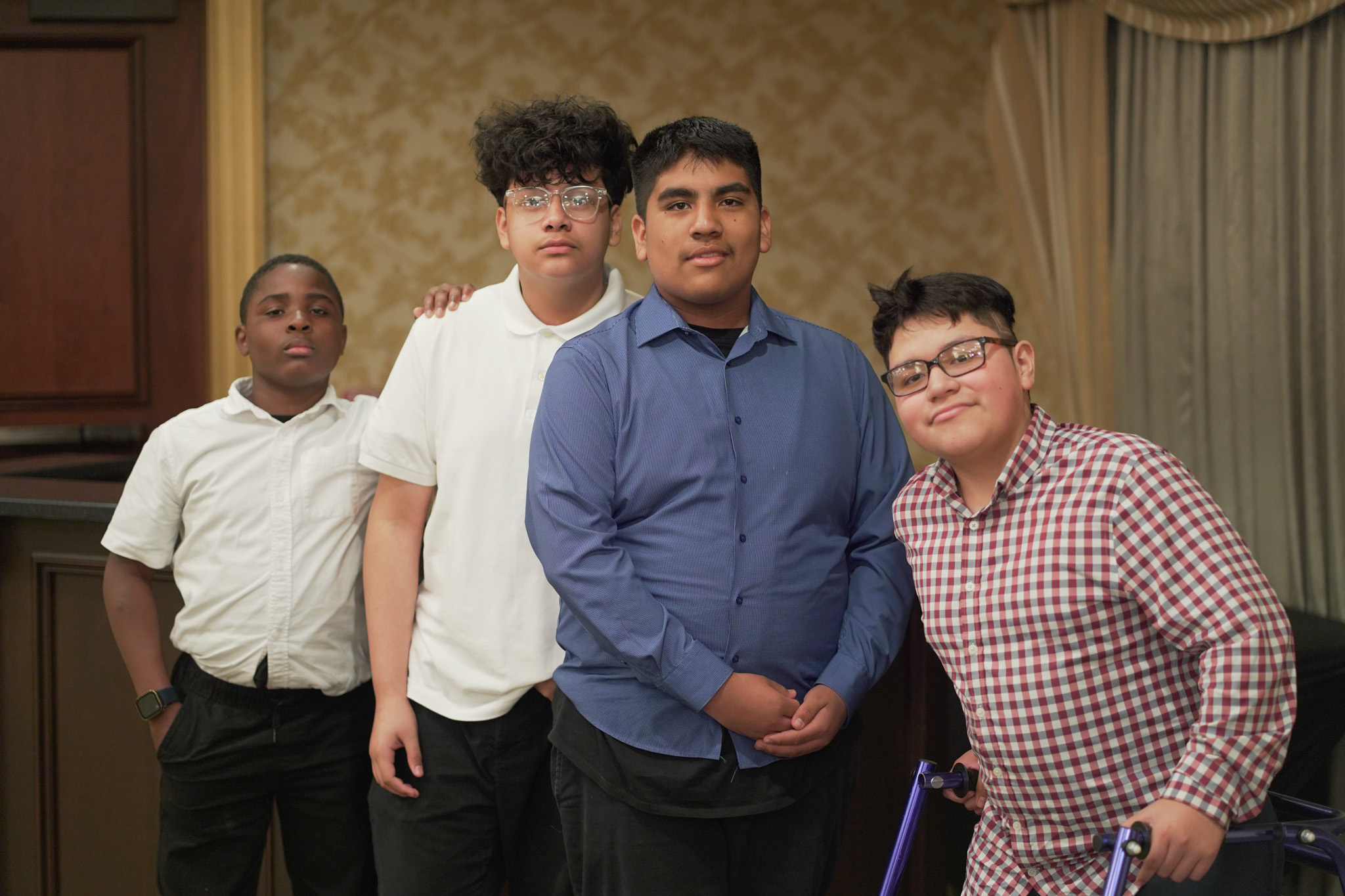 Four boys stand together indoors, posing for a photo. Three wear collared shirts—one blue, two white—while the fourth wears a red plaid shirt and uses a blue walker. A gold patterned wall and draped curtains are in the background.