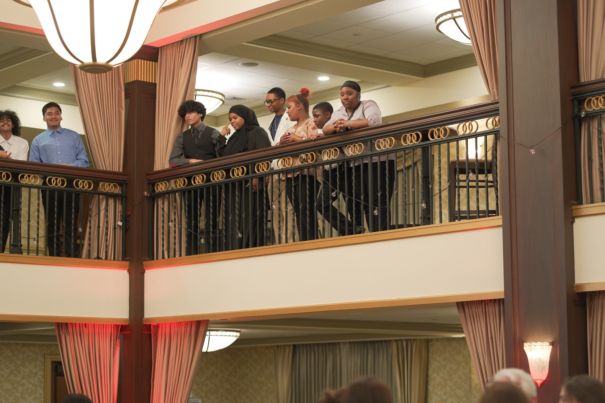 A group of people, mostly young adults, stand and lean on a balcony railing inside an elegant, well-lit venue with chandeliers and curtains, watching something below.